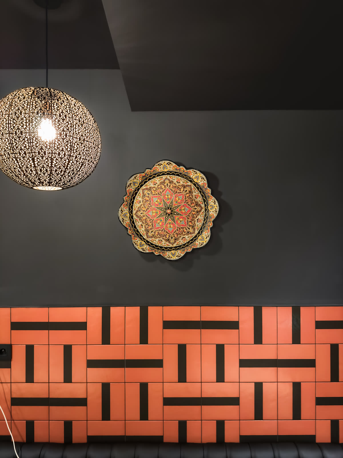 Mosazná lampa a malovaný talíř nad stěnou z terakotových dlaždic